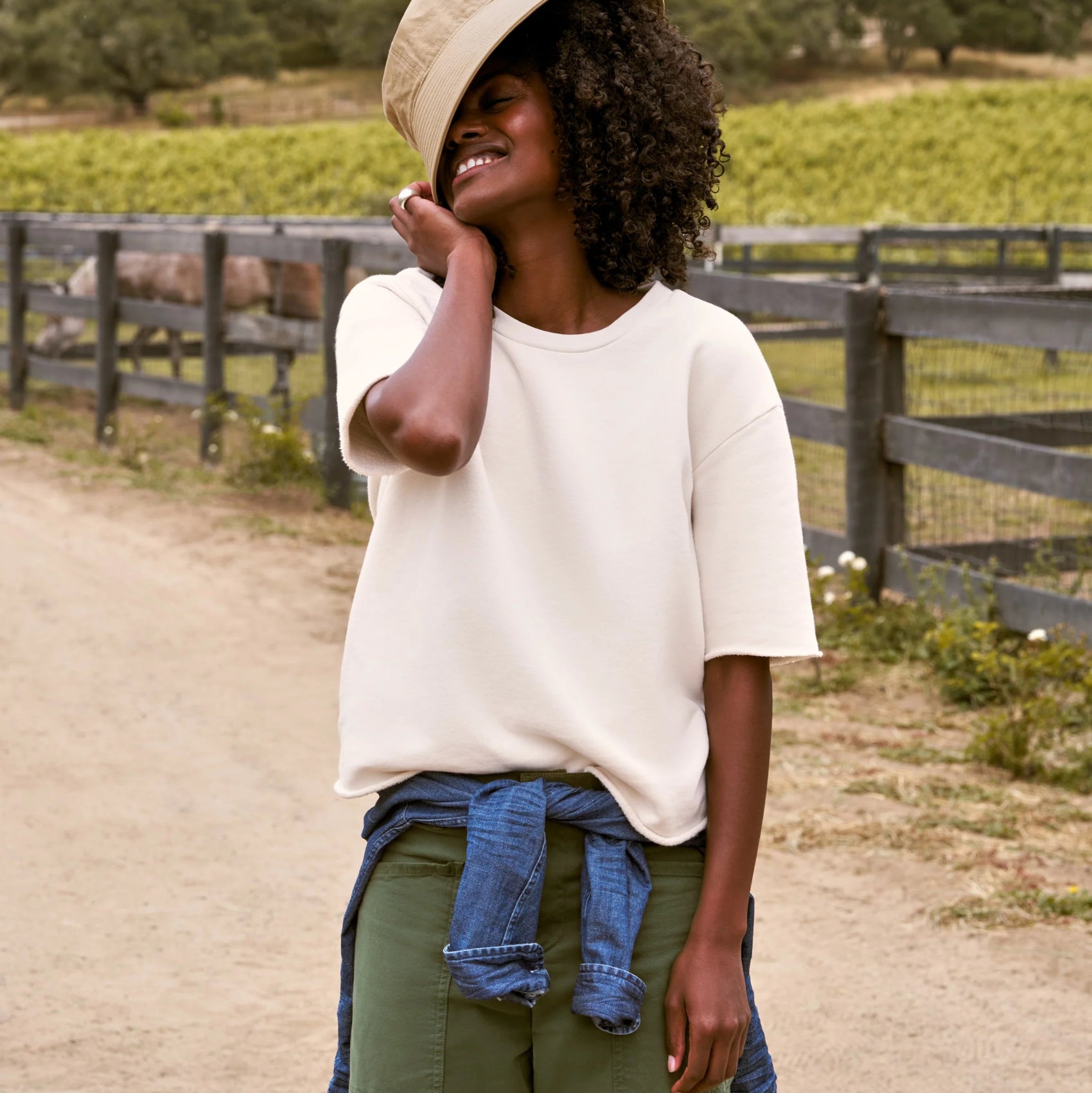 A woman stands on a dirt path wearing an oversized off-white t-shirt, olive green pants, a beige bucket hat, and a blue denim shirt tied around her waist, with a vineyard and wooden fence in the background.